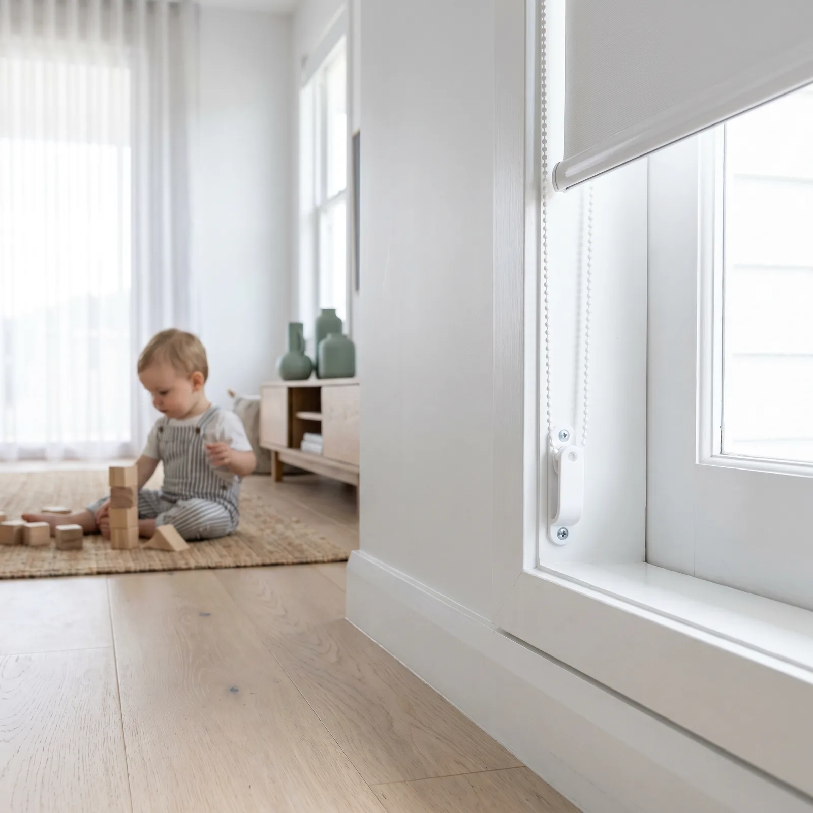Blind chain held taut in a tensioner fixed high on the window frame, a toddler playing safely across the room