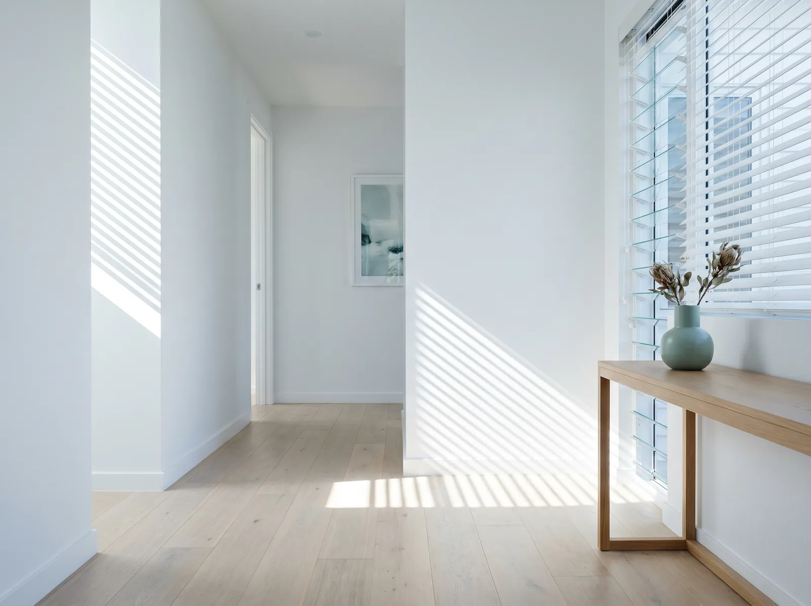 White venetian slats tilted to throw clean parallel bands of light across a hallway wall and floor